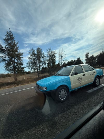 A light blue and white taxi travels on a road, beside trees and a field under a bright, cloudy sky. The car's body is split into two colors: light blue on the bottom and white on the top half. The front license plate is visible, but hard to read. The image is captured at an angle, showing the pavement of another vehicle’s interior.