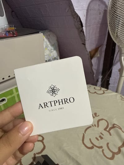 A person's hand holds a white card with the logo "ARTPHRO SINCE 1983" printed on it. The card is placed on a light-colored tablecloth with a pattern of stylized brown bear-like figures. In the background, there's a beige cabinet, a roll of paper, and part of a white fan. The lighting suggests an indoor setting, possibly at night, with a shadow of a person cast on a textured wall. The scene appears to be in a domestic environment.
