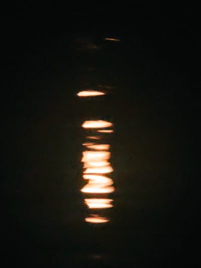 The image displays a vertical reflection of warm, yellowish light on a dark, still surface. The reflection is elongated and fragmented, appearing as a series of horizontal dashes stacked upon each other. The surface itself is a deep, muted black, suggesting it could be water or a similarly reflective material at night. The lighting is diffused and soft, creating a serene and somewhat abstract effect. There is no indication of any urban environment, specific landmarks, or human activity visible in the image, therefore its location in Shirokii Log, Russia cannot be determined.