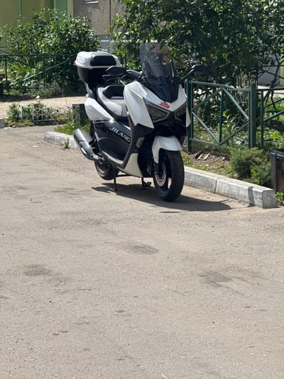 A white and black JILANG scooter is parked on an asphalt surface next to a concrete curb. The scooter has a large black top case and a tinted windshield. Behind the scooter, there is a green metal fence and lush green foliage. In the background, a building with a green facade is partially visible. The scene is illuminated by daylight. The location is the city of Angarsk, Russia.