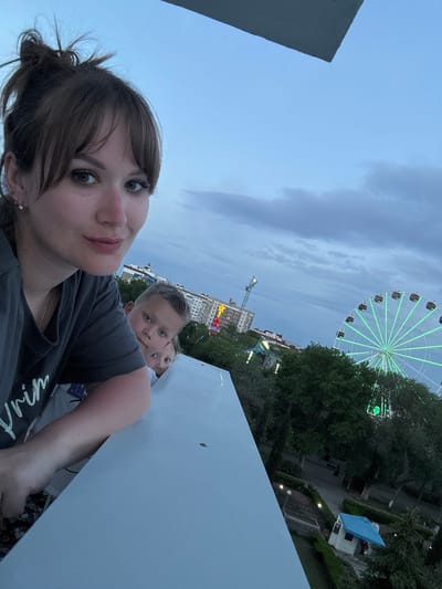 A woman is taking a selfie with two children. The photo includes a ferris wheel with green lights, a crane, a building with lights, and trees. A white structure obscures some of the view. The location is likely Yevpatoriya, Ukraine.
