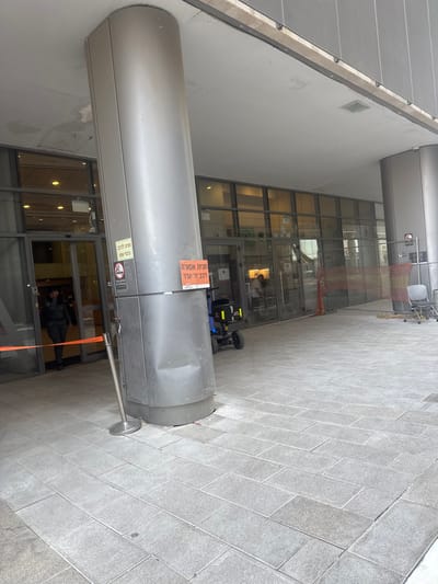 The image depicts the outdoor entrance area of a modern public building in Lod, Israel, characterized by a grey tiled ground and an overhead canopy. Two prominent, silver-toned, cylindrical columns support the canopy; the leftmost column in the foreground exhibits a visible dent on its lower section. Water stains and discoloration are present on the underside of the canopy.

The building's facade consists of multiple floor-to-ceiling glass panels, including automatic sliding glass doors at the entrance. Inside, through the glass, artificial lighting is visible, and two individuals are partially discernible. One person, a security guard or officer in a dark uniform and cap, stands inside near the leftmost set of doors, positioned behind a silver stanchion with an extended orange retractable belt that partially blocks the entrance.

Affixed to the central foreground column are two signs: a yellow "no smoking" sign with graphic symbols and Hebrew text, and an orange rectangular sign with white Hebrew text stating "חניה אסורה" (No Parking) and "רכב דו-גלגלי" (Two-wheeled vehicle). To the right of this column, a small, black and yellow wheeled utility cart is parked. Further to the right, an orange construction mesh barrier partially encloses an area containing a single black chair and other indeterminate objects, adjacent to another supporting column and the building's glass facade. The scene is brightly illuminated by daylight.