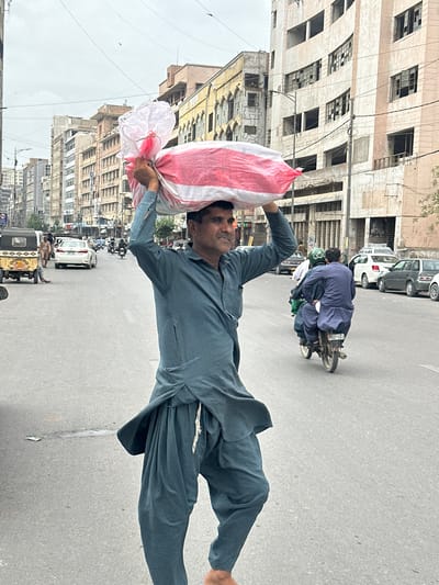 A man in a teal shalwar kameez carries a large, white and red striped bag on his head as he walks across a street in Karachi, Pakistan. He is the central figure in the foreground. Behind him, a motorbike carries two passengers, also in shalwar kameez. Other vehicles, including a white car and a yellow autorickshaw, are visible on the street, along with several pedestrians. The background features multi-story buildings lining the street, some of which appear to be in a state of disrepair. The sky is overcast. The scene depicts a typical urban environment in Karachi.