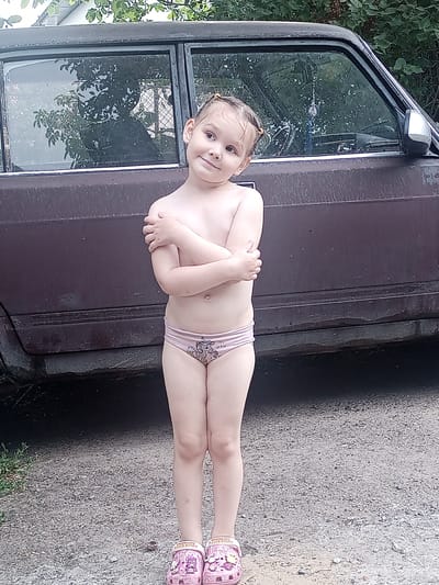 The image shows a young girl standing in front of a dark-colored car. She is wearing pink underwear and pink Crocs with Hello Kitty designs. The girl has her arms crossed in front of her chest and is looking towards the camera with a slight smile. Her hair is pulled back in pigtails, and it appears to be wet. Water droplets are visible on her skin and clothes, suggesting she may have recently been swimming or playing in water.
The setting appears to be outdoors, with a gravel surface on the ground and trees and foliage visible in the background. The car looks old and weathered, with some rust and dirt on its surface. The sky is overcast, indicating a cloudy day.
The image was taken in Yampil, Ukraine. The overall mood of the image is casual and playful, with the young girl appearing comfortable and happy. There is no visible text in the image, except for the Hello Kitty branding on her Crocs.