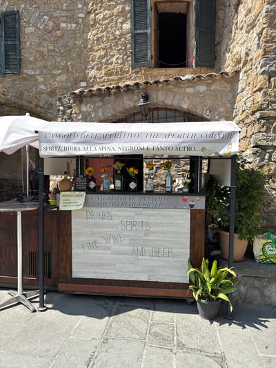 The image captures a small, open-air aperitif stand in the medieval Italian village of Apricale. The stand is set against a rustic stone building, with weathered wooden shutters visible above. The stand itself is covered by a white awning that reads "L'ANGOLO DELL'APERITIVO - THE APERITIF CORNER" in Italian and English, and lists offerings such as "SPRITZ, BIRRA ALLA SPINA, NEGRONI E TANTO ALTRO.....". Below this, a handwritten sign advertises "DRINKS, SPIRITS, FINE WINE AND BEER". The stand displays bottles of alcohol, flowers in vases, and glassware, suggesting it is open for business. Potted plants are visible to the right of the stand, and a table base stands to the left. The ground is paved with cobblestones. The scene is illuminated by natural daylight, casting shadows on the ground. No people are present in the immediate vicinity of the stand.