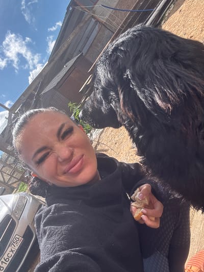 A woman is taking a selfie with a large black dog. The woman is winking and smiling, and she is holding a piece of bread in her hand. The dog is looking at the bread. The background is a rustic outdoor setting, possibly a farm or a rural area. There is a building in the background, and some fencing. The sky is blue with some clouds. The license plate on the car in the background is partially visible and appears to be from Russia. The image quality is sufficient to identify details of the subjects and their surroundings. The location is Dulepovo, Russia.