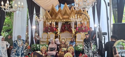 This image depicts a wedding ceremony in Kualacangkoi, Indonesia. A decorated stage with golden accents and floral arrangements serves as the focal point, with ornate chairs suggesting seating for the bride and groom. Several women, some wearing traditional attire and headscarves, are present, with some seated on the stage and others standing in the background. Two young women, also in traditional wedding attire, are seated in the foreground. Men in dark clothing are visible, some operating equipment such as speakers and a fan, indicating a public event with technical support. The overall environment is outdoors, suggested by the greenery visible in the background and the natural lighting.
