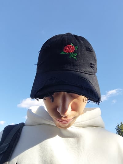 The image displays a close-up, low-angle shot of an individual from the chest upwards, looking downwards. The person is wearing a black, distressed baseball cap featuring a red rose embroidery with a green stem and leaves on the front panel. The brim and crown of the cap exhibit deliberate tears and fraying. The individual wears a white hooded sweatshirt, with a dark backpack strap visible over their left shoulder. Their face is partially obscured by the cap's brim, showing the nose, mouth, and chin. The lighting indicates direct sunlight, casting shadows under the cap and highlighting the person's left side. The background consists predominantly of a clear blue sky with scattered wispy white clouds. A small area of green foliage is visible on the far right side of the frame. The image does not contain specific visual identifiers to confirm the location as Jibb as-Safa, Syria.