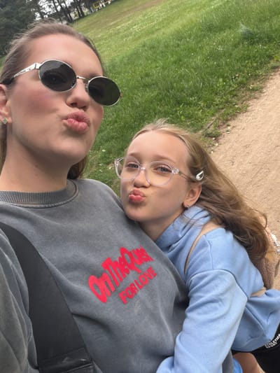 The image is a selfie taken outdoors, likely in a park or grassy area, given the presence of green grass and trees in the background. Two individuals, presumably young females, are the subjects. One, appearing older, is in the foreground, wearing sunglasses and a grey sweatshirt with red text. She is making a kissing face towards the camera. The second, younger individual, is nestled in the arms of the first, wearing a light blue hoodie and clear-rimmed glasses. She is also making a kissing face. Both subjects have blonde hair and appear to be enjoying a casual outing. The lighting suggests it is daytime, with possibly overcast skies or late afternoon light, as there are no harsh shadows. The setting appears to be Ulbroka, Latvia, as indicated by the provided location context. Visible text on the grey sweatshirt reads "Online for Love" in red, stylized lettering.