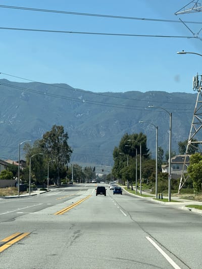 The image depicts a street scene in Fontana, California, USA. A wide, multi-lane asphalt road stretches into the distance, with yellow and white lane markings visible. Several cars are present on the road, with a black SUV in the foreground and other vehicles further ahead. The road is lined with streetlights and mature trees. In the background, a large mountain range dominates the horizon under a clear blue sky. Utility poles and power lines are also visible, as is a tall metal tower on the right side of the image. Residential buildings with pitched roofs are situated along the sides of the road. The overall impression is of a suburban or semi-urban environment with a natural landscape backdrop.