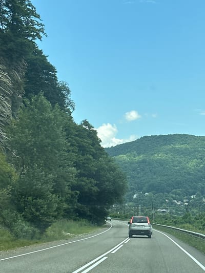 The image depicts a winding road in a mountainous, forested area with a bright blue sky and scattered clouds. A silver SUV with a red stripe on the back is driving on the road, followed by another yellow car in the distance. The road has double white lines indicating two lanes. To the left of the road, a steep, rocky cliff face is covered in lush green trees. To the right, a metal guardrail lines the edge of the road, and beyond that, more trees and a valley with scattered houses and electrical wires are visible. The overall impression is of a scenic drive through a natural landscape. The location of Grecheskii, Russia is not discernable from the image alone.