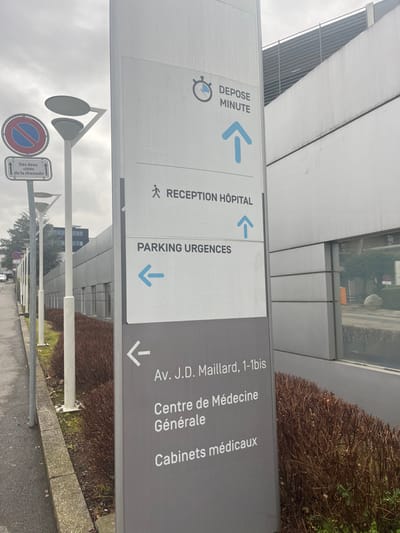 The image shows a tall, vertical directional sign made of two panels, rotated 90 degrees counter-clockwise. The sign is positioned outdoors, likely near a medical facility or hospital, with parts of a modern building visible in the background. The sky is overcast, suggesting daytime with cloudy weather.

The sign serves as a guide for various destinations. The upper, lighter panel, which appears white, indicates:
*   "DEPOSE MINUTE" (Drop-off point/Kiss & Ride) accompanied by a clock icon and an arrow pointing to the left (if the sign were upright, this would be a forward-left direction).
*   "RECEPTION HÔPITAL" (Hospital Reception) with a figure of a person walking and an arrow pointing straight ahead.
*   "PARKING URGENCES" (Emergency Parking) with an arrow pointing to the left (forward-left).

The lower, darker grey panel lists:
*   "Av. J.D. Maillard, 1-1bis" (J.D. Maillard Avenue, number 1-1bis) with an arrow pointing to the right (forward-right).
*   "Centre de Médecine Générale" (General Medical Center).
*   "Cabinets médicaux" (Medical Offices).

In the foreground, there are streetlights and other road signs. One visible road sign is a circular "no stopping or parking" sign (red circle with a red diagonal bar on a blue background). Below it, a rectangular sign reads "Des deux côtés de la chaussée" (On both sides of the roadway), indicating the restriction applies to both sides. Lush, dark green shrubbery borders the area behind the sign and alongside the building. The scene appears to be in Meyrin, Switzerland.