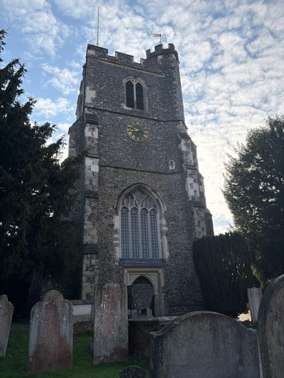 The image displays the tower of St. Augustine's Church, located in Broxbourne, United Kingdom, viewed from its associated graveyard. The prominent church tower is constructed of dark grey, rough-hewn stone, accented with lighter ashlar stone quoins and window dressings, and topped with battlements. A flagpole stands on the left side of the tower's summit, while a weathervane featuring a rooster finial is positioned on the right. A square clock face with gold Roman numerals and hands is embedded into the tower wall, indicating a time of approximately 10:10. Below the clock, a tall, multi-paned Gothic-arched window with intricate tracery is visible, beneath which a smaller pointed arch opening is partially obscured. An upper louvered rectangular opening is located above the clock face. The foreground contains a graveyard with several weathered grey stone grave markers, including upright headstones and a partially obscured table tomb, some displaying reddish-brown discoloration from lichen or moss, all situated on green grass. Large, dark green coniferous trees flank the tower on both sides. The sky above is partially clear blue with scattered white, wispy clouds. No individuals or ongoing activities are present in the scene.