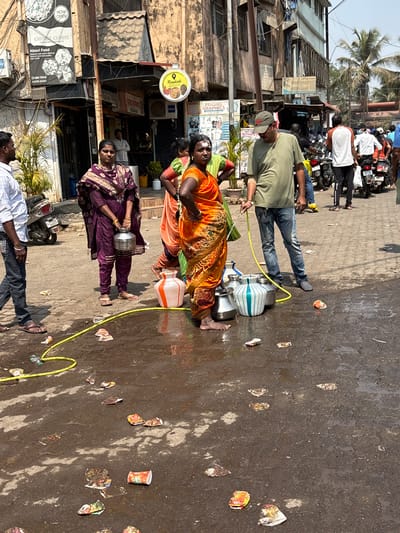 The scene is a busy street in Ambernath, India. The street is littered with discarded food containers and debris. Three women are carrying water containers: two aluminum and one plastic. One woman is wearing a purple sari, another an orange sari, and the third is partially visible. A man in jeans and a green shirt is also carrying water containers. There are shops on the left side of the street, including a Chinese cuisine restaurant and a roadside eatery. Motorbikes are parked along the street, and a crowd of people is visible in the background.