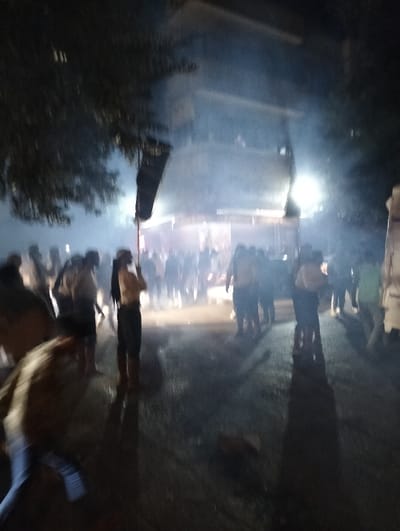 The image depicts a nighttime scene in the city of Homs, Syria, characterized by low light, blur, and atmospheric haze. A crowd of approximately 20-30 individuals is gathered outdoors, predominantly in the mid-ground and background. Artificial light sources illuminate the scene, casting strong beams through visible haze or smoke, and creating bright reflections on the dark, wet ground.
In the mid-ground, slightly to the left of center, an individual stands holding an upright dark-colored flag or banner on a pole. This person wears a light top and dark trousers. Other individuals in the crowd are indistinct due to blur and distance, appearing to be standing or slowly moving. In the immediate foreground, lower left, a person is heavily blurred by motion, suggesting rapid movement or a longer exposure capture of their movement, wearing blue trousers and a light-colored top. Elongated shadows of individuals are cast onto the reflective ground surface.
The background includes a multi-story building, partially visible with some illumination, and silhouetted trees on the left. The overall environment appears to be an urban setting, possibly a street or open area, with a pervasive smoky or foggy atmosphere.