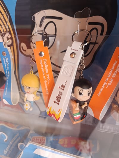 The image shows three keychains hanging in a display case, likely in a retail setting in Noginsk, Russia. The keychains feature cartoon-style figures and branded elements related to Burger King. The keychains are suspended by heart-shaped hooks and metal rings. Each keychain has a character and a rectangular tag with text in Russian and the Burger King logo. The tags also say "Love is...". One tag is blue, one is orange, and one is white. The tags are branded with the phrases "Любовь это... начинать сразу со сладенького" (Love is... start immediately with something sweet), "Любовь это... перекусить по-быстрому" (Love is... to have a quick bite), and "love is..." respectively. A sticker partially visible in the lower area of the image also contains the text "KING" and "BURG" in Russian. The scene is lit and appears to be inside a shop or a similar commercial space.