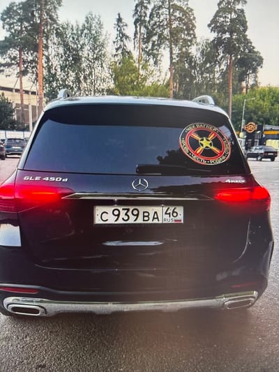 The image shows the rear of a black Mercedes-Benz GLE 450d parked on a wet asphalt surface, likely outdoors in a parking lot. Trees and some buildings are visible in the background. The car has a Russian license plate reading "C 939 BA 46 RUS" and a circular sticker on the rear window. The sticker features a yellow star in the center with red and black elements radiating outwards, containing Cyrillic text. The car's taillights are illuminated red, and the exhaust pipes are visible.