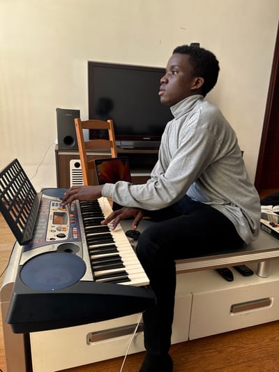 A young man with short, dark hair wearing a gray long-sleeved shirt and black pants plays an electronic keyboard. He is seated on top of a white cabinet with the keyboard placed in front of him. A flat screen TV, wooden stand, and speaker are behind him, with a partially visible laptop. Remotes are also visible on the cabinet near his legs.