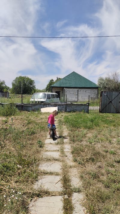 A full-shot, vertical, outdoor, daylight image captures a young girl with blonde hair, wearing a pink long-sleeved shirt and a blue skirt, standing on a paved path with a small bicycle. The girl is facing away from the camera, looking to the right. The path is surrounded by dry grass and weeds. In the background, a white van is parked in front of a building with a green corrugated roof. Further to the right, a corrugated metal gate stands open. The sky above is blue with scattered wispy white clouds, and a thin black wire stretches horizontally across the upper portion of the frame.
