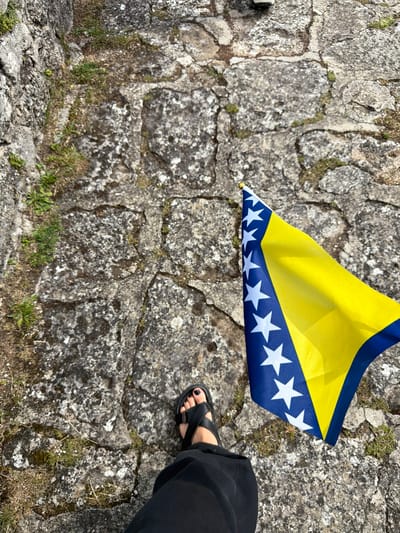 The image shows a person's legs and feet, wearing black pants and black sandals, standing on a stone path. In the foreground, a flag of Bosnia and Herzegovina is held up, displaying its yellow and blue colors with white stars. The background consists of ancient stone blocks, likely part of a historical structure, with patches of grass growing between them. The scene appears to be outdoors, possibly at a historical site. Given the context of Jajce, Bosnia and Herzegovina, this could be the Jajce Fortress or another historical location within the city.