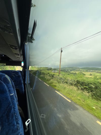 This is a photograph taken from inside a vehicle, possibly a bus, looking out the windshield. The image is oriented sideways, making it appear as if the vehicle is traveling vertically. Outside the windshield, a road curves to the right, bordered by a lush green grassy verge. In the distance, rolling hills covered in fields and dotted with houses suggest a rural landscape. The sky is overcast with a layer of dark clouds, indicating potentially inclement weather, but there are also patches of lighter sky, hinting at the possibility of the sun breaking through.

In the foreground, inside the vehicle, the dashboard and part of a seat are visible. The dashboard appears to be made of dark material, with some reflective surfaces. A blue, patterned seat is visible on the right side of the frame, partially covered by a dark blanket or fabric. A seatbelt buckle is also visible, suggesting that the occupants are secured.

A prominent feature in the middle ground is a wooden utility pole supporting overhead wires. The wires stretch across the frame, disappearing into the hazy sky. The overall impression is that of a journey through a scenic, green, and somewhat moody rural area. The time of day is not definitively clear due to the overcast conditions, but it appears to be daytime. The location is identified as Baltoney Upper, Ireland. There is no visible text in the image.