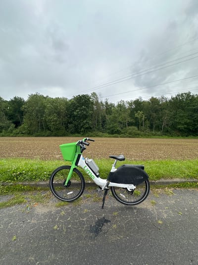 A Lime electric bicycle is parked on the right side of a paved road. To the bicycle's left is a field of recently tilled soil, bordered by a line of trees in the background. Above the trees, the sky is overcast with a layer of gray clouds, and power lines are visible. The bicycle itself is white and green, with a black seat and rear wheel cover. The bike is parked next to the intersection of Rue des Cochets and Rue des Vignes, in Marcoussis, France.