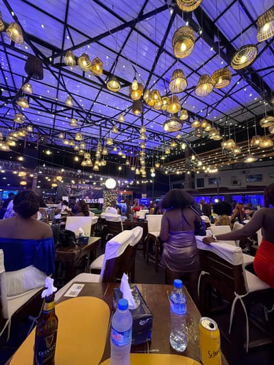 The image depicts an outdoor dining area in Monrovia, Liberia at night. Overhead, a structural framework supports many hanging decorative lights. Tables and chairs are arranged throughout the space, occupied by patrons who appear to be dining and socializing. Bottles of water and beer are visible on the tables, along with napkins and other dining accessories. A banner with the text "It's Party Time!" is visible in the background.