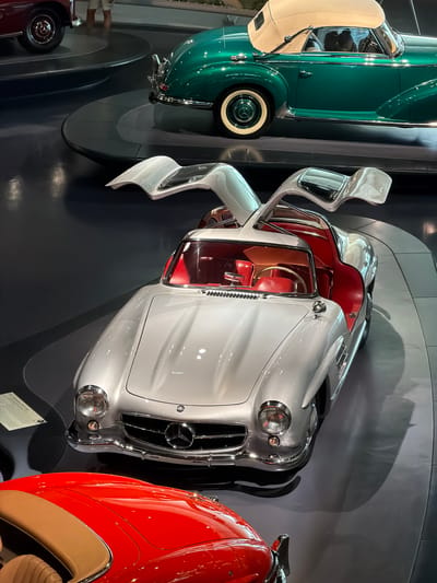 The image displays a collection of vintage Mercedes-Benz automobiles in a museum setting, likely the Mercedes-Benz Museum in Stuttgart, Germany, given the context of vintage Mercedes-Benz vehicles. In the foreground, a silver Mercedes-Benz 300 SL Gullwing is prominently featured with its iconic gullwing doors open, revealing a red interior and steering wheel. To the left and slightly behind it, the front quarter of a red convertible is visible. Further back and to the right, a vintage green Mercedes-Benz convertible with a tan soft-top is parked. A person in light-colored shorts and a dark top is partially visible standing near the rear wheel of a dark red Mercedes-Benz in the upper left corner. The cars are displayed on polished dark gray flooring with curved, raised platforms. The lighting is diffused, casting shadows that highlight the curvature and sleek lines of the vehicles.