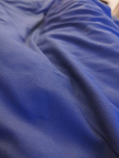 The image is a close-up, shallow-depth-of-field shot of a rumpled, dark blue fabric, possibly a blanket or sheet. The fabric exhibits soft folds and creases, with variations in lighting creating shadows and highlights. There is a single small, dark speck visible on the fabric in the lower center of the image, which could be a dust particle or a minor stain. The background is out of focus and appears to be a lighter, neutral tone. There is no identifiable location in Grohotno, Bulgaria, or any people, objects, actions, or interactions depicted.