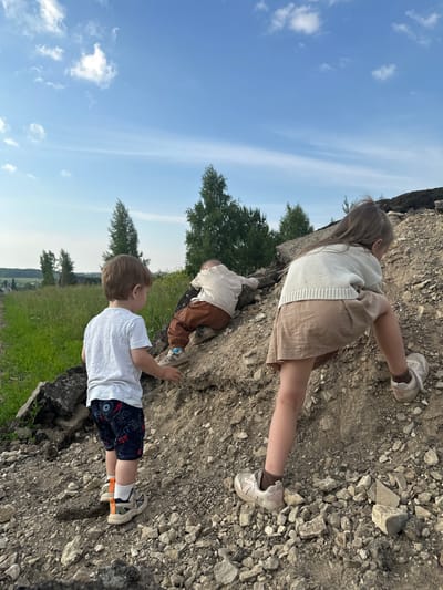 Three children are on a dirt hill under a partly cloudy blue sky. In the foreground, a young boy wearing a white t-shirt and patterned shorts stands looking to his right. Behind him and to his right, a younger child in brown pants and a white shirt is crawling up the hill. To the right of the image, a girl wearing a cream-colored sweater and a beige skirt is climbing the hill, facing away from the camera. The hill is composed of dirt and rocks, with some debris that appears to be asphalt. In the background, there is a green field, trees, and a hint of distant buildings. The setting appears to be a rural or semi-rural area outside of Эстачи, Russia.