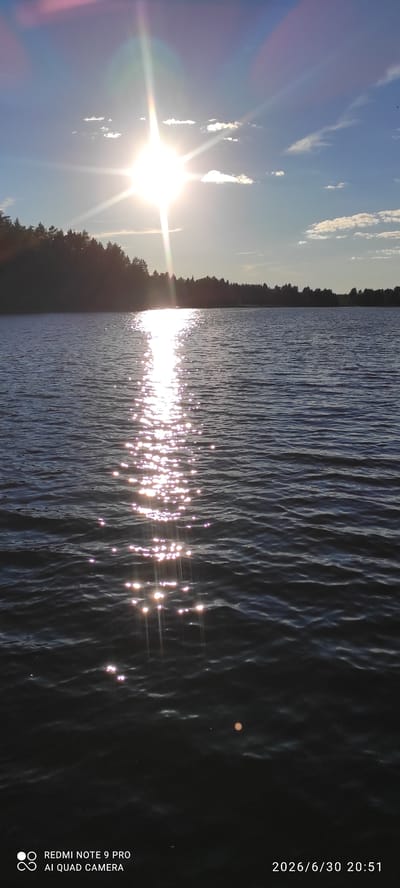 This is a vertical, outdoor, landscape photograph of a body of water, likely a lake, at sunset or sunrise, as indicated by the bright sun low on the horizon and the sparkling reflections on the water. The sun is a brilliant white disc with distinct rays emanating from it, casting a shimmering path across the dark, rippling water. The sky above is a vibrant blue, with scattered white, wispy clouds.
To the left of the water, a dense line of dark green trees forms a treeline, suggesting a wooded shoreline. The light from the sun creates lens flare, with streaks of pink and white visible. The water's surface is textured with many small waves, catching the sunlight to create numerous bright, sparkling highlights that stretch across the scene. In the distance, a small, light-colored circular object is visible on the water, possibly a buoy or a floating object.
The overall mood is serene and peaceful, capturing the natural beauty of a calm evening or morning by the water. The image appears to be taken from a slightly elevated position, looking across the water towards the sun.
Visible text on the right side of the image, oriented vertically, reads: "2026/6/30 20:51" and below that: "REDMI NOTE 9 PRO AI QUAD CAMERA". This text indicates the date and time the photo was taken, along with the device used. The setting is identified as Hartola, Finland.