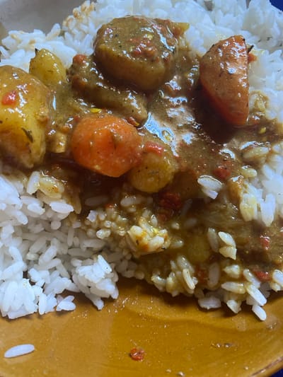 A close-up, overhead view displays a serving of white rice partially covered by a thick, yellowish-brown stew. The stew features various cooked vegetables, including rounded orange carrot pieces, lighter yellow potato chunks, and a distinct reddish-orange wedge resembling a tomato. Numerous small red and dark specks, likely spices or chili flakes, are visible throughout the stew. The cooked white rice grains are clearly distinct beneath and around the stew. The meal rests on a plain, mustard-yellow colored plate. This scene is located in Mape, Nigeria.