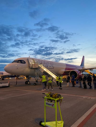 This image captures a Wizz Air Airbus aircraft parked on an airport tarmac, likely at dusk or dawn, indicated by the warm, low-angle sunlight and the atmospheric sky. The sky transitions from a clear, lighter blue near the horizon to a deeper blue with scattered cumulus clouds and some darker, more defined cloud formations higher up.

The primary activity is the boarding or deboarding of passengers. A line of individuals, some carrying bags, is seen ascending a set of mobile stairs to enter the aircraft's front left door. Several ground crew personnel, easily identified by their high-visibility yellow vests, are present on the tarmac and near the stairs, overseeing the operations.

Notable details include the distinctive purple and white livery of the Wizz Air aircraft, with "Wizz Air" clearly legible on its fuselage. The mobile boarding stairs display text such as "BER High Efficiency Stands" and the identifier "A07." On the tarmac, ground markings with numbers like "072" and painted white and red lines are visible. To the right, various pieces of ground support equipment are present, including a bright yellow unit bearing the "swissport" and "Goldhofer" logos.

The overall scene suggests a routine airport operation. Given the "BER" text on the stairs and the provided context, the location is Berlin Brandenburg Airport in Schönefeld, Germany.