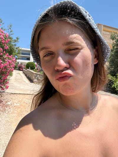 The image is a selfie taken outdoors on a sunny day, likely in Grad Šibenik, Croatia, given the presence of Mediterranean flora and architecture. A young woman, wearing a light blue crocheted hat and a thin silver necklace, is the primary subject. Her face is turned towards the camera, with one eye squinted shut and her lips pursed in a playful expression. The background reveals a paved pathway leading uphill, bordered by a low stone wall and lush pink flowering bushes. Behind the path, a modern white building with balconies is visible, along with mature trees and clear blue sky. The lighting suggests it is midday, casting distinct shadows on the woman's skin. The overall scene depicts a casual, holiday atmosphere.