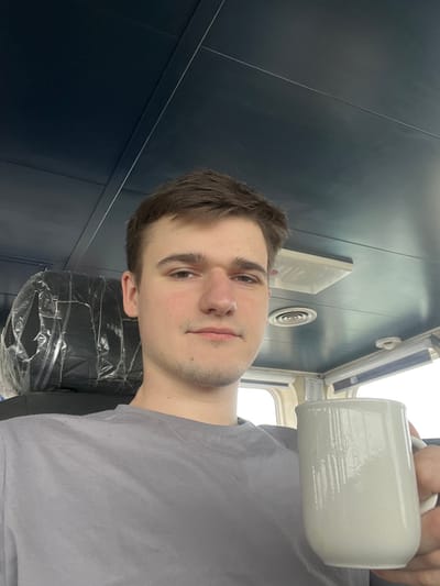 A young adult male, with short brown hair and light facial stubble, wears a grey t-shirt and looks directly at the camera with a neutral expression. He holds a white ceramic mug in his right hand, positioned to the right of the frame. He is seated indoors within what appears to be a vessel's or vehicle's cabin or bridge. The interior features a dark blue paneled ceiling with white strips and a square light fixture, along with a circular vent. Behind him, a seat with a clear plastic cover over the headrest and upper back is visible. Windows line the background to the right, showing an indistinct, bright exterior, suggesting daylight conditions. The specific location within Bangor, United Kingdom, cannot be identified from the visual evidence present in the image.