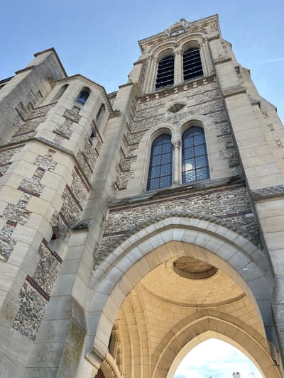 The image shows a low-angle view of the facade of a stone church in Rambouillet, France, likely Église Saint-Lubin. The architecture features a mix of large, light-colored stone blocks and rougher, darker stonework with interspersed red bricks. The building has arched windows, some with stained glass, and a prominent bell tower with arched openings and a clock face near the top. The sky above is clear and blue. The focus is on the architectural details and textures of the church.