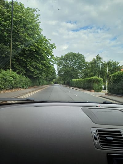 The image is a first-person view from inside a car driving on a two-lane road lined with trees and hedges. The road has a tarmac surface with white lane markings. The sky is overcast with patches of blue sky visible. There is a lamppost on the left side of the road and houses can be seen in the distance on the right. The car's dashboard is visible in the foreground. There are no people or animals visible in the scene. The location is identified as Middlesbrough, United Kingdom.