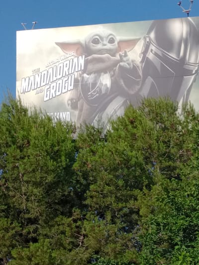 This media file is an image of a billboard featuring characters from the Star Wars franchise. The billboard depicts The Mandalorian, a helmeted figure, and Grogu, a young alien with large ears and eyes. The title "THE MANDALORIAN AND GROGU" is prominently displayed in large, stylized font, along with "STAR WARS." The image on the billboard is faded and appears to be an advertisement for the show.
The billboard is positioned outdoors, with a clear blue sky visible in the background. To the right of the billboard, lush green foliage from trees partially obscures the advertisement, suggesting an outdoor setting with vegetation. There are also some small, unidentifiable objects attached to poles in the lower left corner of the image, which could be part of a lighting system or signage. The image is taken from a low angle, looking up at the billboard. The time of day appears to be daytime, given the bright sky. There are no specific weather cues, but the scene suggests fair weather. The presence of a large billboard advertising a popular franchise implies a public space, possibly a roadside or an urban area. The overall mood is informative, showcasing promotional material for a known entertainment property.
