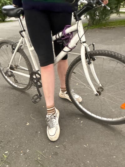 A person is standing next to a white bicycle on an asphalt surface, possibly in a park or on a street. The person is wearing black leggings, a multi-colored long-sleeved shirt or sweater, white sneakers with grey laces, and striped socks. A purple bicycle lock is attached to the frame of the bicycle. The bicycle has a black seat, a chain, pedals, and wheels with tires. The background shows trees and foliage, suggesting an outdoor setting. The image does not provide any specific information about the location in the city of Nevyansk, Russia.