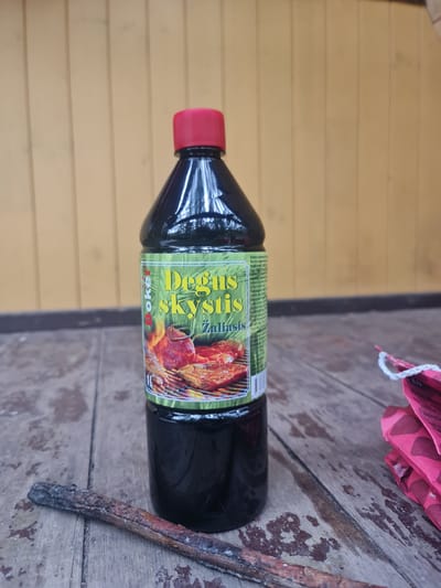 The image shows a bottle of "Degusi skystis" (burning liquid) with a red cap on a wooden surface. The bottle has a label with text in Lithuanian and an image of meat grilling. To the left of the bottle is a burnt stick, and to the right is a pink object with a white string. The background consists of yellow wooden planks. The scene appears to be outdoors, likely on a porch or patio, in Stanuliškės, Lithuania, given the context. There are no people or visible actions occurring in the image.