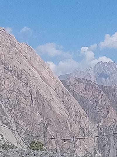 The image displays a panoramic view of a rugged mountain range under a bright blue sky with scattered white clouds. The mountains are large, imposing, and rocky, with visible textures and geological formations. In the foreground, a dark, thin wire stretches horizontally across the frame, obscuring a portion of the lower mountain slopes. The ground at the very bottom appears to be covered in loose gravel or small stones. A small patch of green vegetation is visible on the left side of the frame, near the base of the mountains. The scene is a natural landscape with no discernible human activity or structures, suggesting a remote or undeveloped area. It is plausible this view is from the vicinity of Zeravshan-2, Tajikistan, known for its mountainous terrain.