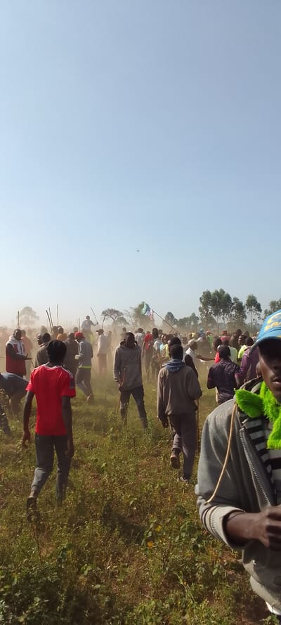 The image captures a large gathering of people, predominantly men, in a dusty, grassy field in Isukha ICHINA, Kenya, under a bright, hazy blue sky. A significant amount of dust is visible in the air, particularly in the middle ground, indicating active movement by the crowd.
The subjects are numerous individuals, many of whom are carrying long, slender sticks or clubs. They are spread across the field, with some in the foreground, others in the midground partially obscured by dust, and more in the background near a line of trees. In the immediate foreground to the right, a person is partially visible, wearing a blue cap with an unreadable white logo and a distinctive bright green, feathery neck accessory. Another person in the foreground, seen from behind, wears a red t-shirt.
The scene is an open, rural or semi-rural landscape. The ground is covered with green and brown vegetation. In the distant background, scattered trees with light green foliage are visible. The sky is clear but appears very hazy, especially closer to the horizon, contributing to the dusty atmosphere. A very small, dark, indistinct object can be faintly observed high in the sky.
The activity appears to be a communal gathering or movement, possibly a march, protest, or a traditional event, given the presence of sticks carried by many. The time of day is clearly daylight, likely late morning or early afternoon due to the bright sun. No clear, readable text is discernible on any objects or clothing. The overall impression is one of a dynamic and possibly intense outdoor event.