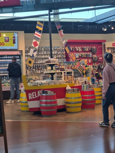 The photograph depicts an indoor retail environment, likely a gift shop, in Moreira, Portugal. A prominent display in the center of the frame is shaped like a boat, constructed with red and white striped components, and flanked by yellow and red barrels. This display bears the word "RELAX" vertically. A banner of signal flags is strung across the display, adding to the nautical theme.

Merchandise, including souvenirs, trinkets, and packaged goods, is densely arranged on shelves behind the boat display. Customers are present: one individual is browsing the refrigerated section below the display, while another is seen from above near the back of the shot. The flooring is polished and reflects light. A refrigerated case is visible on the left side of the frame, displaying beverages with a price tag of "50€". Above the cooler is advertising for "Coca-Cola" and "Frito," as well as text "IN WELLNESS CLASS. STAY HYDRATED." Overhead lighting illuminates the scene. The image is rotated ninety degrees clockwise.