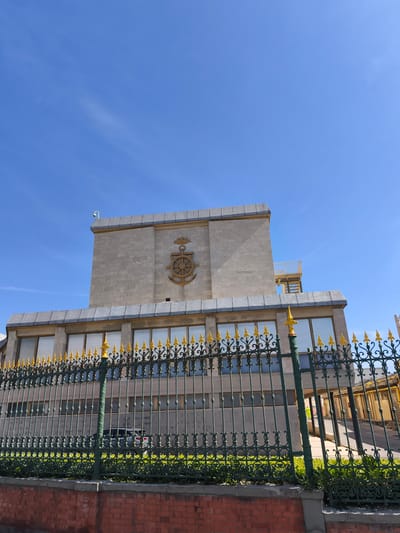 The image depicts the exterior of a building in Livorno, Italy, identified as the "Palazzo dell'Accademia Navale". The building features a light stone facade with a prominent emblem of an anchor and crown on its upper section. A dark green metal fence with gold decorative elements runs along the front of the property, separating it from a brick wall in the foreground. Through the fence, a portion of the building's lower level with reflective windows is visible, as well as a glimpse of a car and some greenery. The sky is a clear, bright blue with a few faint white clouds. A security camera is mounted on the upper corner of the building.