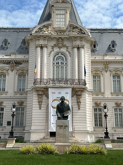 The image depicts the facade of the National Art Museum in Craiova, Romania. The building is of classical architectural style, with ornate detailing, including columns, windows with decorative surrounds, and a roof with prominent dormer windows. A banner advertising an exhibition is hung on the facade beside a bronze statue of a person. The sky is blue with scattered clouds. A grassy area and plantings can be seen in the foreground. A lamp post is visible to the right.