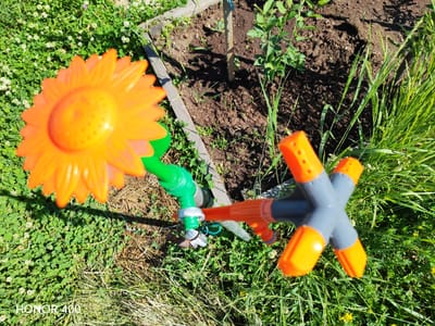 The image shows a garden sprinkler shaped like a sunflower, in the city of Erkhirik, Russia. The sprinkler is connected to a green hose and is positioned on a patch of green grass with small white clover flowers. Behind the sprinkler, a garden bed with dark soil and young plants is visible, bordered by wooden planks. The sunlight is casting shadows on the ground and plants. The "HONOR 400" watermark is visible in the lower left corner of the image.