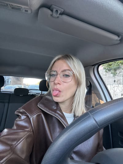A woman sits in a car, wearing a brown leather jacket and glasses. Her blonde hair is styled straight. She sticks her tongue out towards the camera in a playful manner. A steering wheel is visible, along with the car's interior, including the sun visor. Outside the car window, buildings are seen.