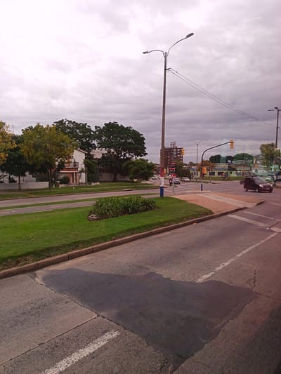 The image depicts a street scene in Montevideo, Uruguay, under an overcast sky. The foreground shows a paved street with visible patching and a white lane marking. A median strip separates the street, featuring a manicured lawn and a small bed of greenery. In the midground, there is a residential area with houses, trees in varying stages of foliage, and a pedestrian crossing with a striped pattern. Further back, there are buildings, including a taller structure that could be an apartment complex. Streetlights and traffic signals are visible at an intersection where several cars are present. A dark van is prominent on the right, approaching the intersection. A person is seen walking near the intersection on the left. The overall scene suggests a typical urban or suburban neighborhood with traffic and pedestrian activity.