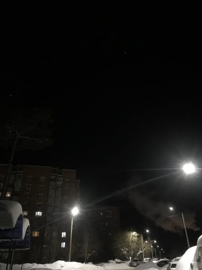 At night, a tall apartment building with lit windows stands next to a dark building in the background. Street lights illuminate the scene, their bright beams cutting through the darkness and revealing snow-covered surfaces. Bare trees can be seen near the buildings. Some clouds are visible above the buildings.