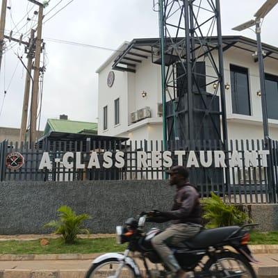 The image shows the exterior of "A-CLASS RESTAURANT" in Kabala, Nigeria. The restaurant building is white with dark trim and large windows. A black metal fence in front of the building displays the restaurant's name in large white letters and a circular logo with cutlery. A man on a motorcycle is blurred as he rides past in the foreground. Overhead power lines and poles are visible to the left, and a green-roofed building is partially visible behind the restaurant. The sky is overcast.